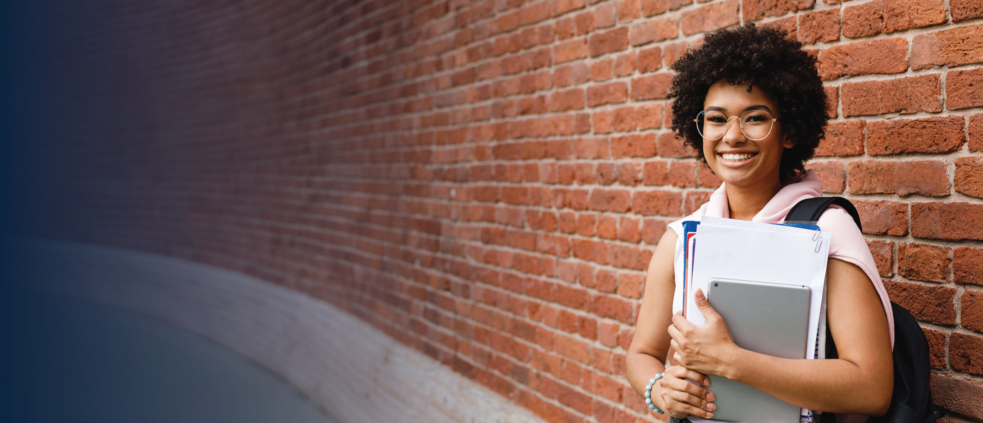 Female student standing against a brick wall holding books and a tablet.