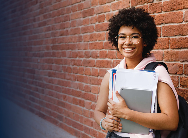 Female student standing against a brick wall holding books and a tablet.